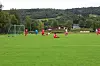 Fotballspillere i røde og blå drakter spiller på en grønn bane med mål og skogkledd bakgrunn.
