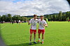 To fotballspillere står på en grønn bane med andre spillere og trær i bakgrunnen.