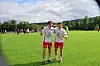 To fotballspillere står på en grønn bane med andre spillere og trær i bakgrunnen.