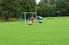 To fotballspillere ligger på gresset foran et mål på en bane med skog og en bil i bakgrunnen.