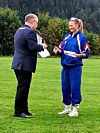 To personer står på en gressbane og holder papirer mens de snakker utendørs.