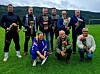 En gruppe på åtte personer poserer på en fotballbane med blomsterbuketter og fjell i bakgrunnen.