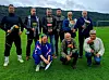 En gruppe på åtte personer poserer på en fotballbane med blomsterbuketter og fjell i bakgrunnen.