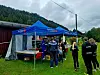 Folk står ved et blått Speider-telt på en gresslette med skog og fjell i bakgrunnen.