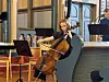En cellist spiller i et kirkerom mens andre musikere og notestativer står bak.