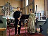 To musikere står foran noter og instrumenter i en kirke med rødt gulv og altertavle i bakgrunnen.
