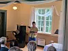 Barn leser fra en bok i en stue med piano, stort vindu og publikum i forgrunnen.