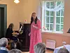 Kvinne i rosa kjole synger ved piano i et lyst rom med publikum i forgrunnen.