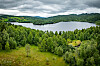 Landskap med innsjø, tett grønn skog og åser under mørke skyer.