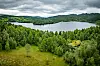 Landskap med innsjø, tett grønn skog og åser under mørke skyer.