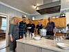 Fem personer står samlet i et kjøkken med store gryter, glass og serveringsutstyr på en kjøkkenøy.