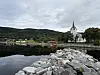 Utsikt over en steinlagt strand mot en liten havn, hvit kirke og grønne fjell under grå himmel.