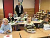 Barn sitter ved pulter i et klasserom, mens en mann med ordførerkjede står i bakgrunnen.