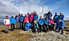 En gruppe mennesker står samlet på en steinete fjelltopp med blå himmel og sjø i bakgrunnen.