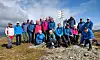 En gruppe mennesker står samlet på en steinete fjelltopp med blå himmel og sjø i bakgrunnen.