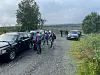 Vandrere med ryggsekker går på en grusvei ved parkerte biler i et grønt, overskyet landskap.