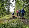 Turgåere i jakker går over en smal trebru ved et vann omgitt av skog.