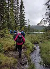 Turgåere med sekk går langs en smal sti ved en bekk i en skog.