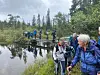 Turgåere går over en smal trebro ved et rolig vann i et skogsområde.