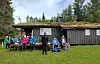 Gruppe turgåere sitter og står utenfor en hytte med torvtak i en grønn skoglysning.