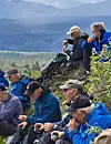 Flere turgåere sitter på en gress- og steinskråning med fjellandskap i bakgrunnen.