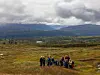 Turgåere samlet på en gresskledd fjellhei med dal, skog og fjell under skyet himmel.