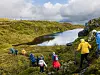 Turgåere i fargerike jakker går langs en sti ved en elv i et grønt fjellområde.