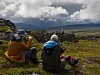 To personer sitter på bakken i et fjellområde med sekk og kamera mens mørke skyer ligger over landskapet.