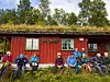 Turgåere sitter foran en rød hytte med torvtak i skogkanten.