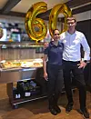 To personer står sammen ved en bar, omgitt av gullballonger med tallet 60.