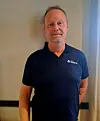 Portrett av en smilende mann i mørk poloskjorte foran en lys vegg.
