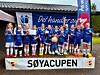 Et jentefortballag i blå fotballdrakter står samlet med pokal foran et klubbhus.