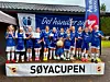 Et jentefortballag i blå fotballdrakter står samlet med pokal foran et klubbhus.
