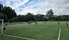 Fotballspillere i kamp på en kunstgressbane omgitt av trær og tilskuere.
