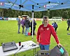 Mann i rød jakke ved et utstyrsbord på en fotballbane under en blå teltduk.