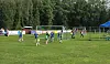 Unge fotballspillere i aksjon på en gressbane med mål, trær og tilskuere.