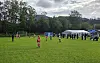 Barn spiller fotball på en gressbane med telt, tilskuere og skog i bakgrunnen.