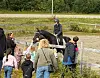 Rytter på mørk hest foran barn og voksne ved et inngjerdet rideområde.