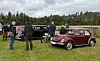 Rød veteranbil, flere klassiske kjøretøy og besøkende på et grønt område.