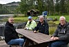 Fire menn sitter rundt et trebord ved en skogkledd vannkant.