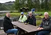 Fire menn sitter rundt et trebord ved en skogkledd vannkant.