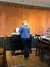 Person spiller piano foran en trevegg i et rustikt rom med bord og stoler.