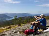 To personer sitter på et fjell med ryggsekker og ser ut over en innsjø og fjell under blå himmel.