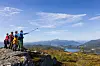 Gruppe turgåere på en fjellknaus med utsikt over fjord, åser og fjell under blå himmel.
