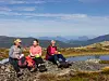 Tre fotturister hviler ved et fjellvann i steinete fjellterreng.