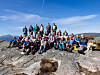 Stor gruppe turgåere samlet på et steinete fjellplatå under blå himmel.
