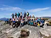 Stor gruppe turgåere samlet på et steinete fjellplatå under blå himmel.