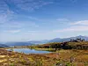 Fjellvann omgitt av lyng og steiner, med vandrere på høydedraget under en blå himmel.