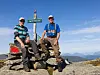 To turgåere står ved et toppskilt på et steinete fjell med fjellandskap i bakgrunnen.