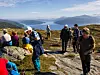 Turgåere på en steinete fjellsti med innsjø og fjell i bakgrunnen.